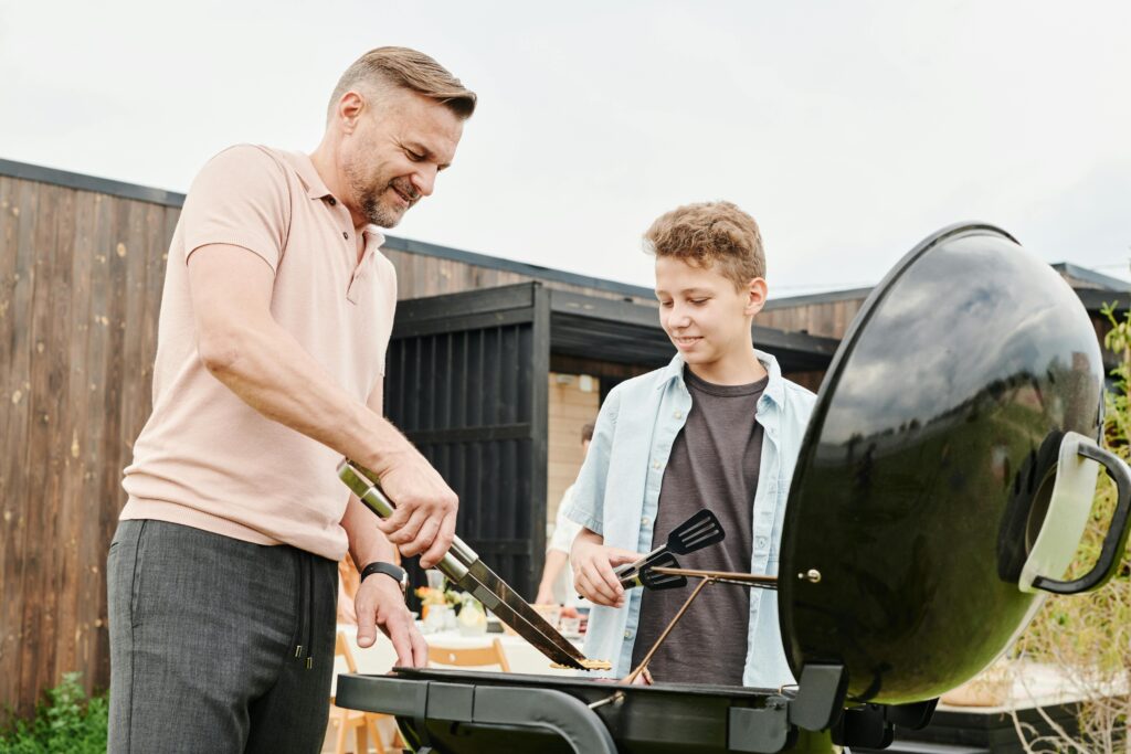 Father and son bonding while cooking on a grill outdoors. Sunny day family barbecue.