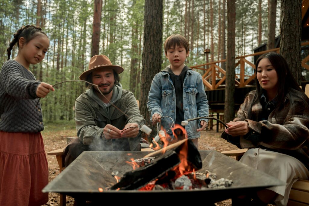 A diverse family roasting marshmallows over a campfire on a sunny day in the woods.